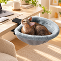 Tabby cat curled up in a grey plush Brighty DeskNest attached to a wooden desk beside a laptop, showing the raised edges and black clamp attachment.
